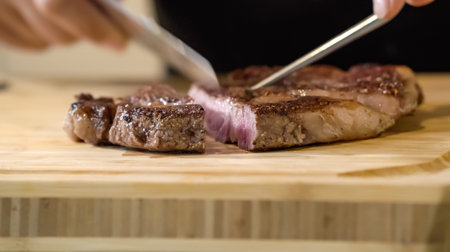 The close up image of a chef cutting medium rare grilled beef steak with chef knives on a wooden...