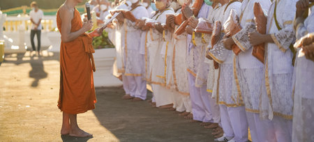 Participants in white clothing line up holding folded orange robes as soft sunlight reveals intricate lace details and hands within a medium to large ceremonial setting.