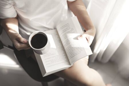 Young women reading a book and holding cup of coffee sitting at window enjoys of rest, Girl reading a book.の写真素材