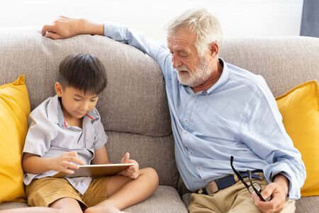Grandfather and little cute grandson playing touchscreen tablet PC together on sofa in their houseの写真素材