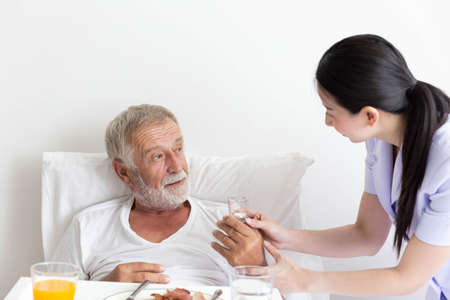 Nurse give medicine pills to elderly senior patient while he is in his bed with breakfast