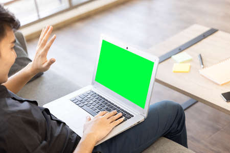 A young man greeting his friend online while using his computer to have an online conference with green screen during the work from home period because of pandemicの写真素材
