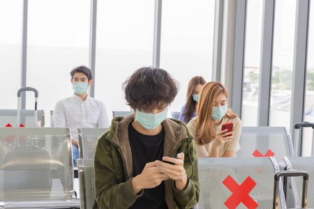 A group of young Asian passengers sit and wait in an airport terminal for their flight with masks on...