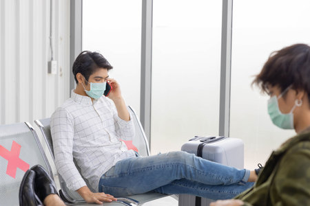 An Asian man tourist with suitcase luggage wearing surgical mask talking with his smartphone sitting...