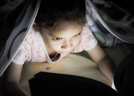 A little cute girl reading on the bed at nightの写真素材