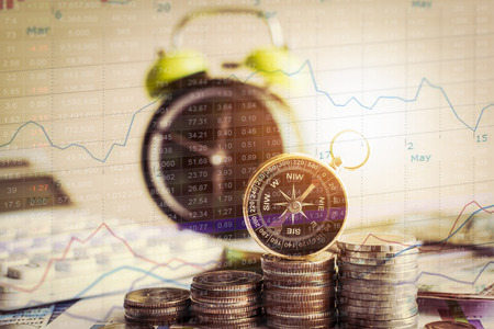 Close up compass on pile of coins with alarm clock and stock market graph information. Investment...
