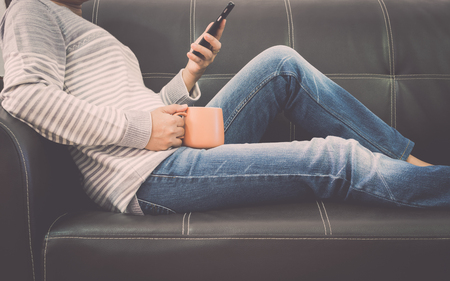 Woman is on leather sofa using mobile phone and holding a coffee cup at home.の写真素材