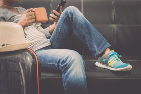 Female traveller is relaxing on leather sofa holding mobile phone and coffee cup with traveling bag at the airport.Travel concept.の写真素材