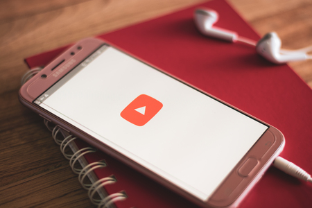 PHUKET, THAILAND - AUGUST 23, 2018 : Smartphone Samsung Galaxy J7 pro open apps youtube application on the screen with earphones and notebook on wooden table.のeditorial素材