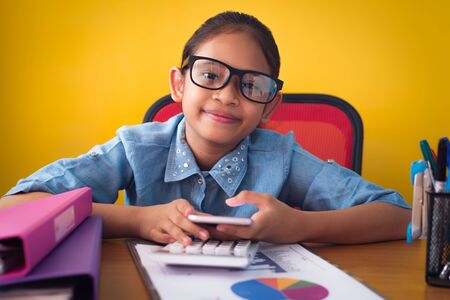 Cute girl smiling and holding a mobile phone on working desk isolated yellow background, Education concept.の写真素材