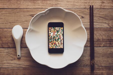 Mobile phone with food picture on screen in a plate on wooden table with chopsticks and spoon, Technology consumption.の写真素材