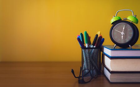 Alarm clock, spectacles, color pens and stack of books are on wooden table isolated yellow background for education equipment concept.の写真素材