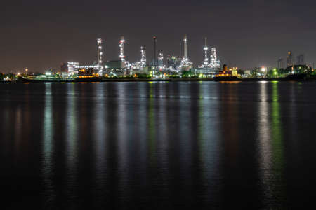 Night photo crude oil refinery plant and many chimney with petrochemical tanker or cargo ship at coast of the river with colorful bright light from lamp reflect on water at thailandの写真素材
