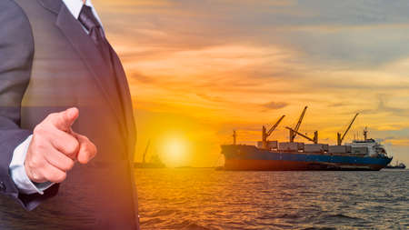 business man wearing a suit point finger forward on image Large cargo ship for transportation import export goods while anchor at sea in evening with golden sunset backgroundの写真素材