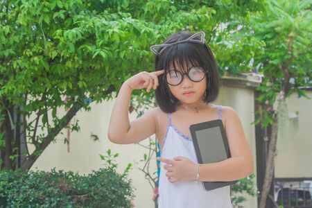 Cute little Asian Chinese girl in glasses holding smartphone and thinking in the garden.の写真素材