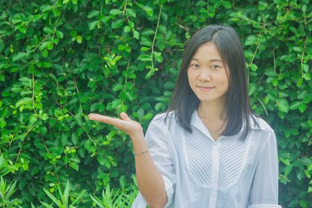 Shoot photo Asian woman portrait wear white shirt, holding open hand and smiling with green tree background. (Autumn filter effect)の写真素材