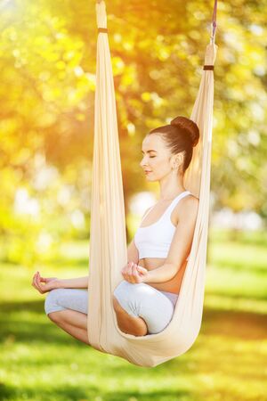 Anti-gravity Yoga outdoor. Happy woman doing yoga exercises, meditate in sunny park. Yoga meditation in hammock. Concept of healthy lifestyle and relaxation. Pretty woman relax at yoga hammockの写真素材