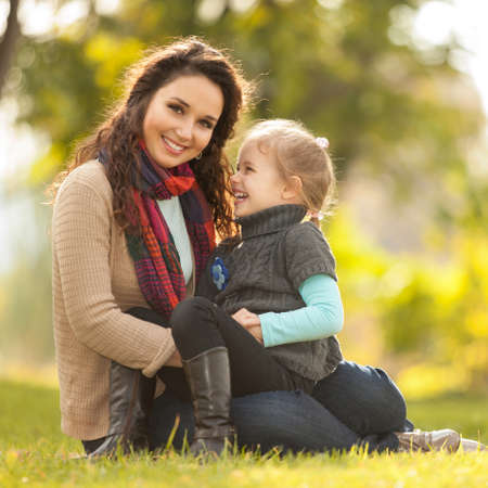 Mother and daughter walking in the autumn park. Beauty nature scene with colorful background, yellow trees and leaves at fall season. Autumn outdoor lifestyle. Happy family relax on green grassの写真素材