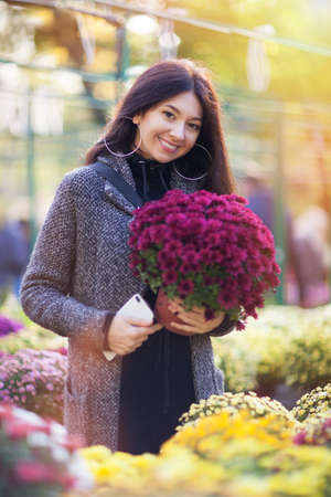 Young pretty woman buying flowers at outdoor market or shop. Beauty nature scene with colorful floral garden background, yellow and pink flowers at fall season. Autumn outdoor lifestyleの写真素材