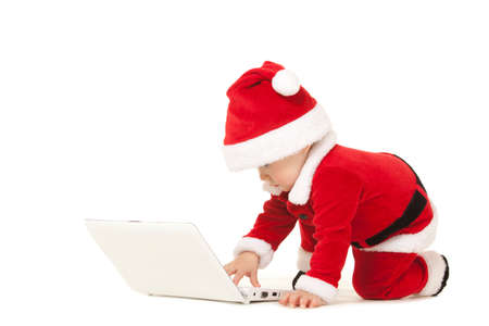 Cute santa baby on the white background. Small model in santa hat at studio holding white laptop. Christmas, xmas, winter concept. Santa girl looking at white laptopの写真素材