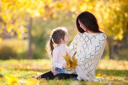 Happy mother and daughter in the autumn park. Beauty nature scene with family outdoor lifestyle. Happy family resting together on green grass, having fun outdoor. Happiness and harmony in family lifeの写真素材