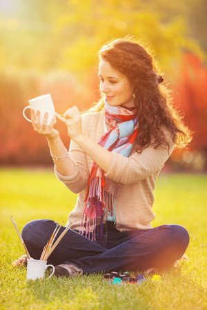 Pretty woman paints cups in the autumn park. Beauty nature scene with colorful foliage background, yellow trees and leaves at fall season. Autumn outdoor lifestyleの写真素材