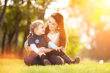 Mother and daughter walking in the autumn park. Beauty nature scene with colorful background, yellow trees and leaves at fall season. Autumn outdoor lifestyle. Happy family relax on green grassの写真素材