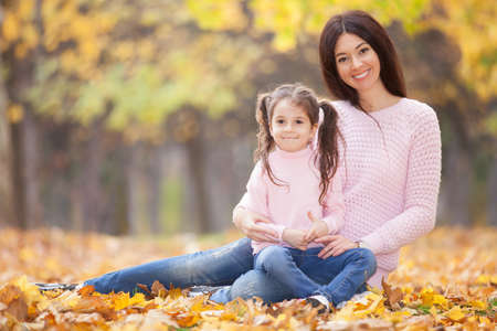 Happy mother and daughter in the autumn park. Beauty nature scene with family outdoor lifestyle. Happy family resting together on green grass, having fun outdoor. Happiness and harmony in family lifeの写真素材