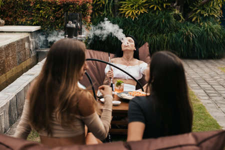 Group of young women with dark hair smoking a hookah and exhaling white smoke.の写真素材