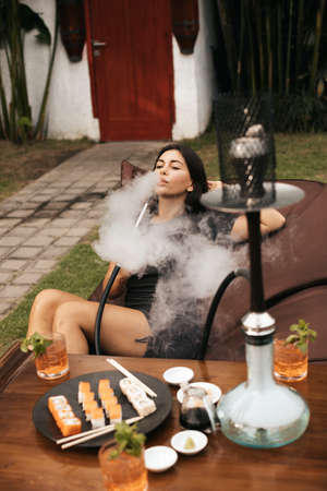 Young European woman with black hair in grey dress smokes hookah or shishaの写真素材
