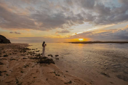 Bride in a long white wedding dress stands on the on Bali ocean beach at sunsetの写真素材
