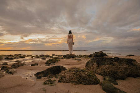 Bride in a long white wedding dress stands on the on Bali ocean beach at sunsetの写真素材