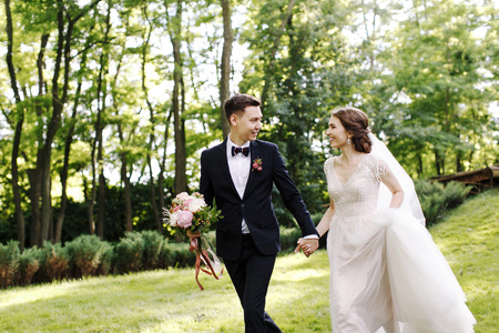 Happy smile bride and groom look at each other and running in the green garden. Wedding in the summer in the park. Happy wedding couple. Stylish and beautiful. Princesss dress.の写真素材