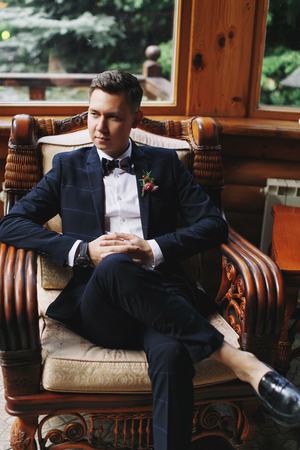 stylish groom portrait on background of wooden wall. rustic wedding concept in barn, happy bohemian newlyweds. man relaxing in vintage chairの写真素材