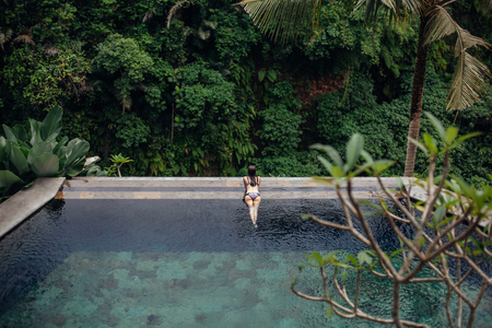 Slim sexy brunette woman in swimsuit relaxing on edge tropical infinity pool in jungle. Palms around, crystal clean water. Luxury resort on Bali island.の写真素材