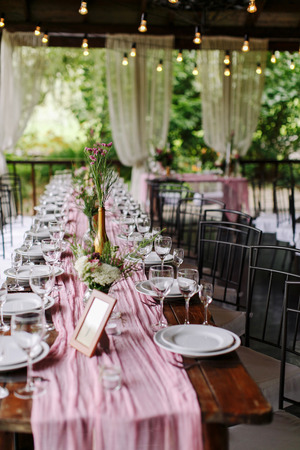 Wedding. Banquet. The festive table for guests, decorated with a composition of white and pink flowers and greenery, there are candles, covered with the tablecloth in wooden houseの写真素材
