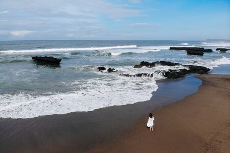 Aerial view of unrecognizable woman in white dress walking near ocean and enjoy summer day on beach....