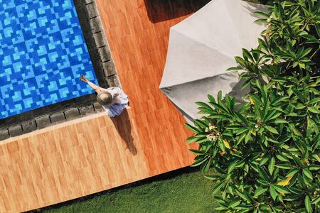 Top view of young unrecognizable woman in white dress and straw hat sitting near swimming pool....