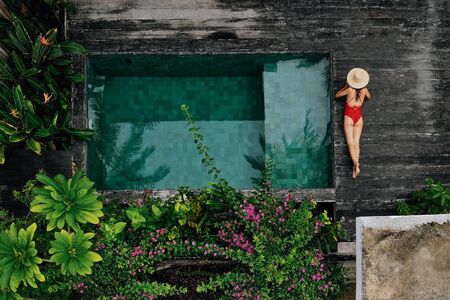 Enjoying suntan. Vacation concept. Top view of slim young unrecognizable woman in red bikini and straw hat lying near swimming pool in jungle. Back view, without face.の写真素材