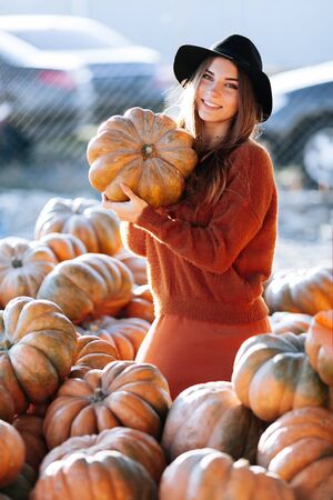 Portrait of happy woman with ripe orange pumpkin in hands on background of farmers market in brown sweater and hat. Cozy autumn vibes Halloween, Thanksgiving day.の写真素材