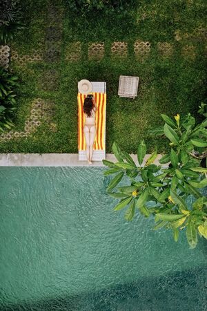 Top view of unrecognizable slim young woman in beige bikini relaxing and sunbathe near luxury swimming pool in green tropic in Ubud.Vacation concept. Drone photoの写真素材