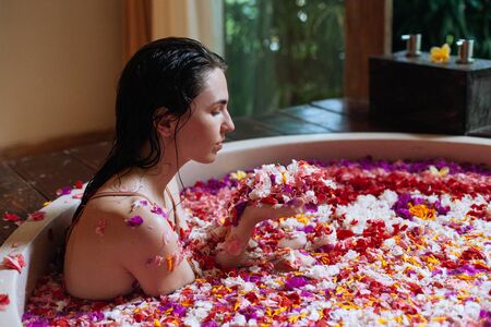 Beautiful young woman holding petals in hands in outdoor spa. Luxury round bath tub with jungle view. Natural organic tropical petals in the water: white, pink, red, violet. Beauty treatment conceptの写真素材