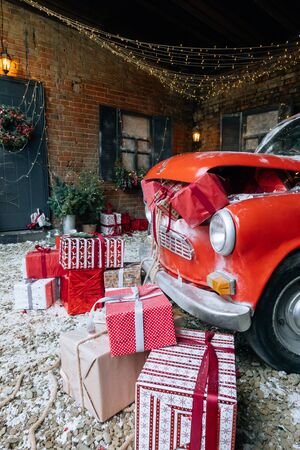 Christmas gifts packed with red,white,craft paper and tapes under decorated retro red car with trunk full of New Year gifts on snow and festive lights and garlands outside of houseの写真素材