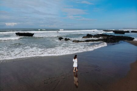 Aerial view of unrecognizable woman in white dress walking near ocean and enjoy summer day on beach....