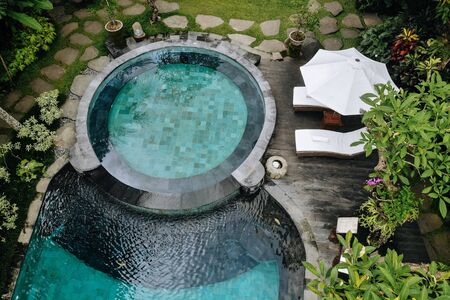 Aerial view of luxury round pool with white umbrellas and sun beds in tropical jungle and palm...