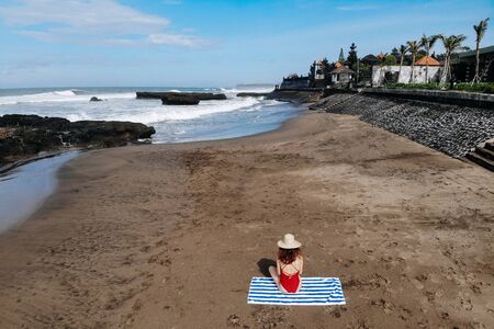 Aerial view of woman in red bikini and straw hat sitting and getting tanned on towel on ocean beach with black sand. Vacation in Bali. Photo from droneの写真素材