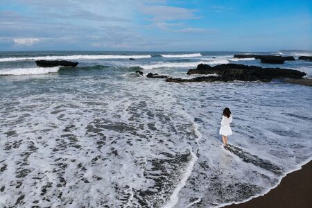Aerial view of unrecognizable woman in white dress looking at ocean and enjoy summer day on beach with black sand. Vacation in Bali. Photo from drone. Copy space. Photo from droneの写真素材