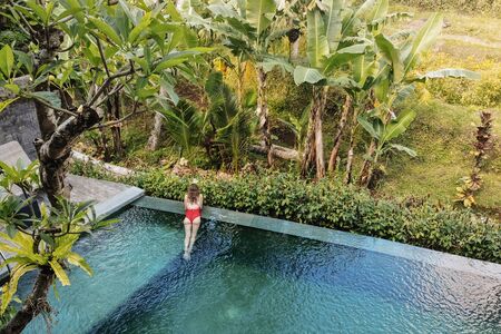 Girl in red bikini in a private pool in Bali admires a beautiful view of the palm trees.Luxury holiday.Woman resting in the pool with a beautiful view of the palm trees on the island of Bali.の写真素材
