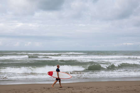 CANGGU, BALI, INDONESIA - FEBRUARY 25, 2019: young surfer man walking on beach with surfboard at summer on coast of Bali island. Recreation and water sport during vacations in Indonesia.のeditorial素材
