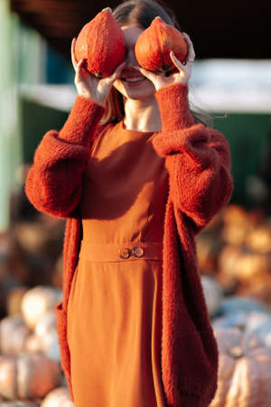 Portrait of happy woman with ripe orange pumpkins in hands close eyes near wagon with orange pumpkin on farmers market in brown sweater, dress. Cozy autumn vibes Halloween, Thanksgiving dayの写真素材